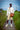 A model stands on a path outdoors wearing a cream sweatshirt, beige shorts with green patterns, white socks, and cream sneakers, while carrying a brown leather shoulder bag. The background shows a grassy field, water, and a lighthouse under a blue sky.