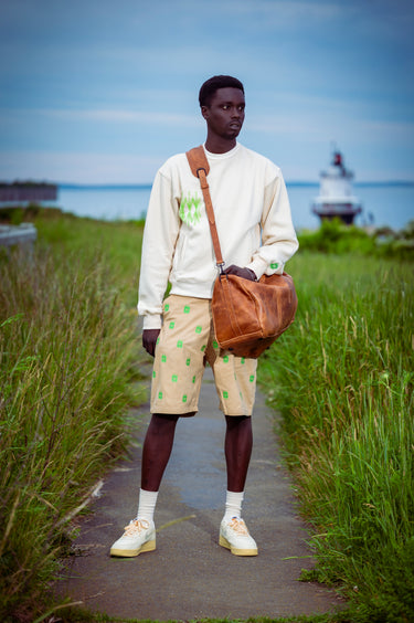 A model stands on a path outdoors wearing a cream sweatshirt, beige shorts with green patterns, white socks, and cream sneakers, while carrying a brown leather shoulder bag. The background shows a grassy field, water, and a lighthouse under a blue sky.