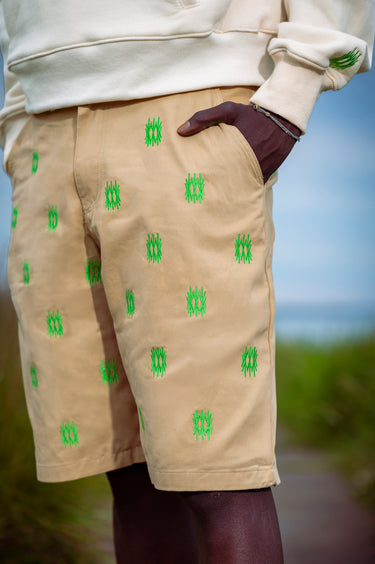 Beige shorts with a repeating green embroidered logo pattern, worn by a model with a cream sweatshirt and dark tights, set against a blurred outdoor background.