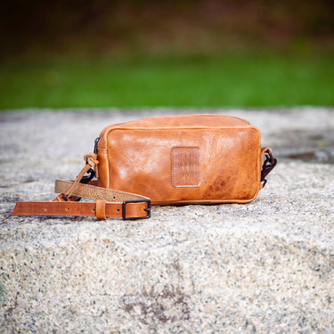 A tan leather crossbody bag with a matching leather strap, resting on a textured stone surface with a blurred green and brown outdoor background.
