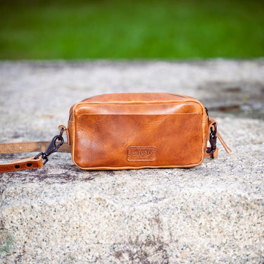 A small tan leather crossbody bag with a rectangular shape and a visible 'LUXE' logo patch, resting on a textured stone surface with a blurred green background.