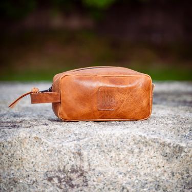 A small tan leather pouch or cosmetic bag with a zipper closure and a small rectangular patch on the front, resting on a textured stone surface with a blurred outdoor background.