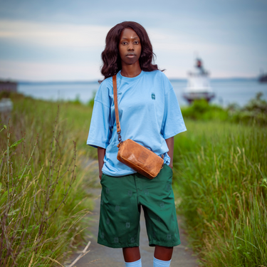 A person standing on a path outdoors wearing a light blue oversized T-shirt, green shorts, light blue socks, and green sneakers, with a brown shoulder bag. Tall grass lines both sides, and water and a distant lighthouse are visible in the background.