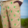 Beige shorts with a repeating green embroidered logo pattern, worn by a model with a cream sweatshirt and dark tights, set against a blurred outdoor background.