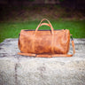 A brown leather duffel bag with top handles and a shoulder strap, resting on a large gray stone outdoors with grass and trees blurred in the background.