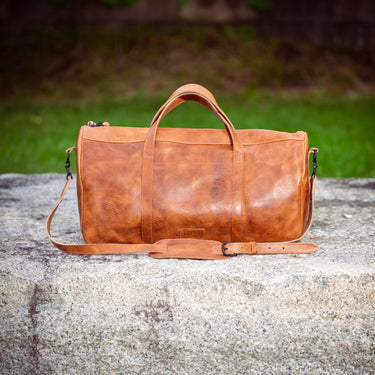 A brown leather duffel bag with top handles and a shoulder strap, resting on a large gray stone with a blurred green outdoor background.