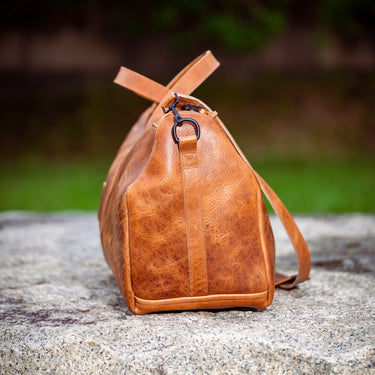 A brown textured leather duffel bag with a matching shoulder strap, resting on a stone surface with a blurred green outdoor background.