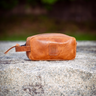 A small tan leather pouch or cosmetic bag with a zipper closure and a small rectangular patch on the front, resting on a textured stone surface with a blurred outdoor background.